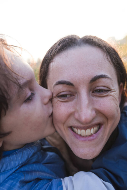 Un niño le da una flor a su madre, un niño besa y abraza a mamá, una mujer huele una flor, un retrato de una mujer sonriente, una maternidad feliz - Foto, Imagen