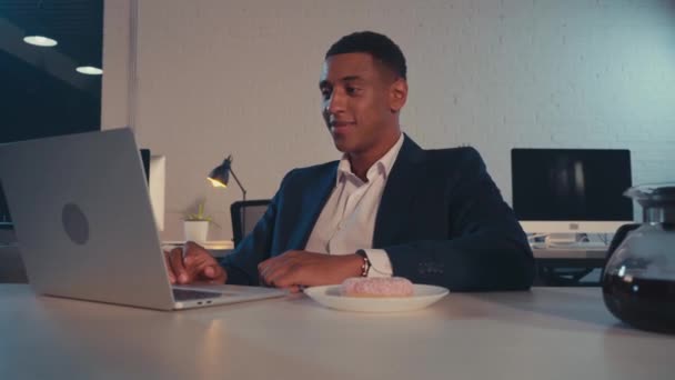 szczęśliwy afrykański amerykański biznesmen jedzenie pączka w pobliżu laptopa w biurze - Materiał filmowy, wideo