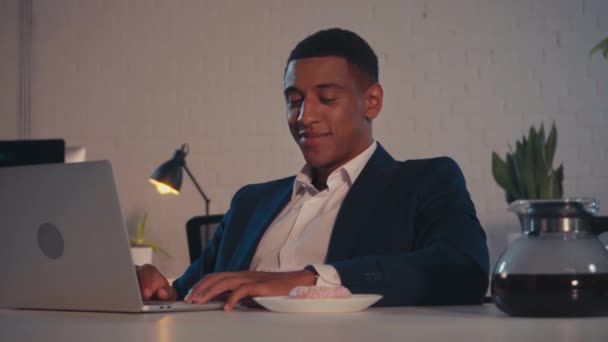 felice uomo d'affari africano americano sfregamento mani e mangiare ciambella in ufficio - Filmati, video