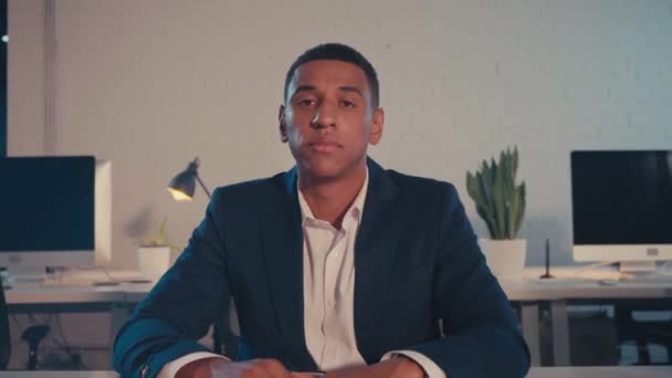 νεαρός Αφροαμερικάνος επιχειρηματίας που κοιτάζει κάμερα στο γραφείο του τη νύχτα - Πλάνα, βίντεο