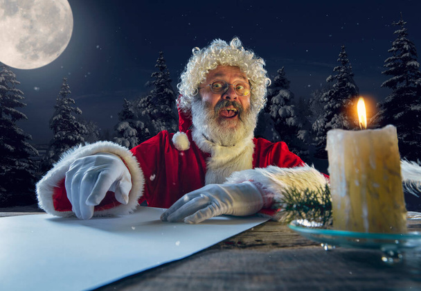 Emotional Santa Claus congratulating with New Year and Christmas, writing a letter, wish list in midnight with candle - Fotó, kép