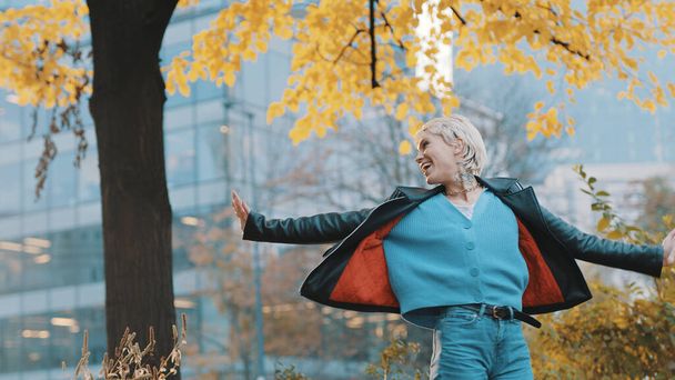 Junge glückliche kaukasische Frau in Lederjacke mit ausgestreckten Händen, die sich an einem Herbsttag unter dem Baum im Park dreht - Foto, Bild