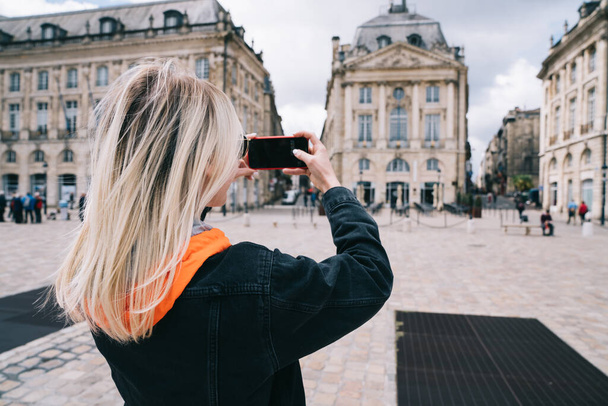 Back view of young woman in black jeans jacket enjoying architecture and taking picture with smartphone during exciting journey in France - Фото, зображення