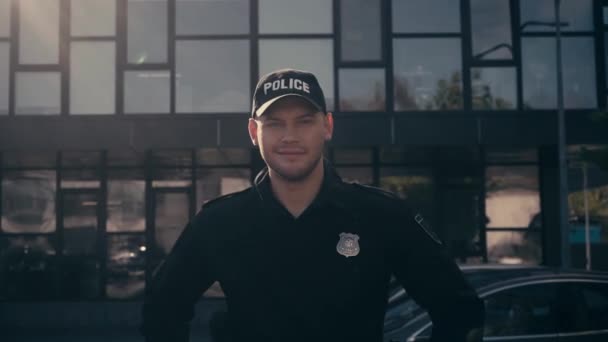 Glimlachende politieman kijkt naar camera in de buurt van auto 's en gebouw buiten  - Video