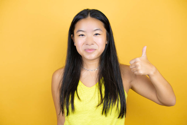 Jeune belle femme asiatique sur fond jaune isolé souriant et faisant le signal ok avec son pouce - Photo, image