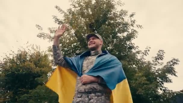 Glimlachende soldaat zwaaiend met de hand terwijl hij de Oekraïense vlag vasthoudt en buiten loopt  - Video