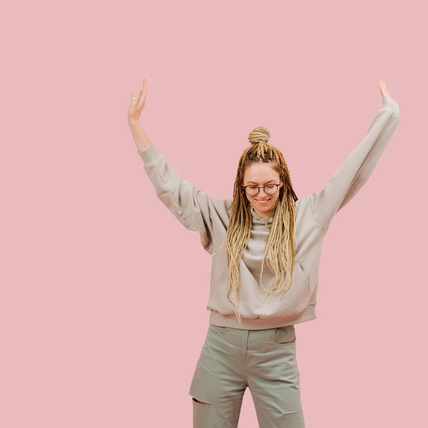 Happy dancing woman in round glasses with stylish blond afro braids over pink background. She,s looking down and smiling, hands outstreched up. - Фото, изображение