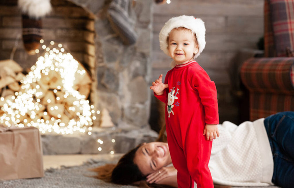 Klein meisje in kerstmankostuum kijkt in de camera in de buurt van de kerstverlichting en mama lacht naast haar. Nieuwjaarsavond - Foto, afbeelding