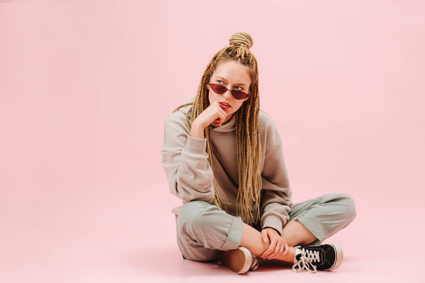 Bored young woman in narrow sunglasses with stylish blond afro braids sitting cross-legged on the floor over pink background. Wearing loose grey clothes and sneakers. - Foto, Bild