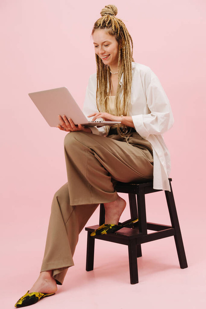 Cheerful young woman with stylish blond afro braids sitting on a stepping stool with laptop in hands over pink background. She's smiling, looking at the screen. - Fotoğraf, Görsel