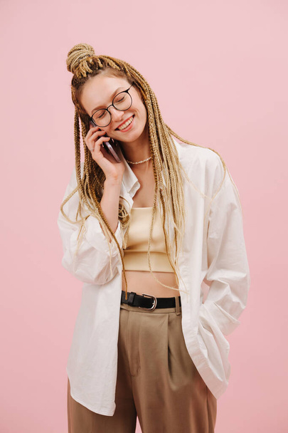 Happy young woman in narrow sunglasses with stylish blond afro braids talking on phone with her eyes closed. Over pink background. - Photo, Image