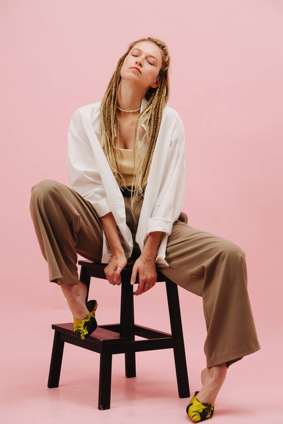 Provocative stylish young woman with stylish blond afro braids sitting on a stepping stool over pink background. Legs spread, pointing toes in shoes, eyes closed. - Fotografie, Obrázek