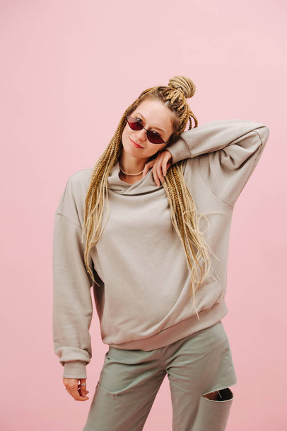 Pretty smiling young woman in narrow sunglasses with stylish blond afro braids over pink background. Playing cute, posing, shifting weight on one leg. - Фото, изображение