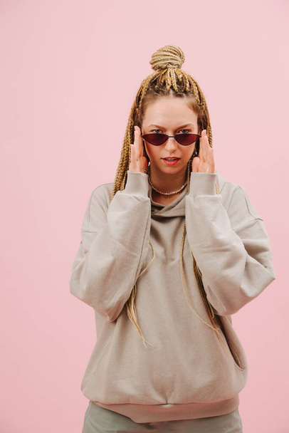 Sensual provocative young woman in narrow sunglasses with stylish blond afro braids over pink background. Posing, looking over sunglasses, lowering them with palms of her hands. - Foto, Imagen