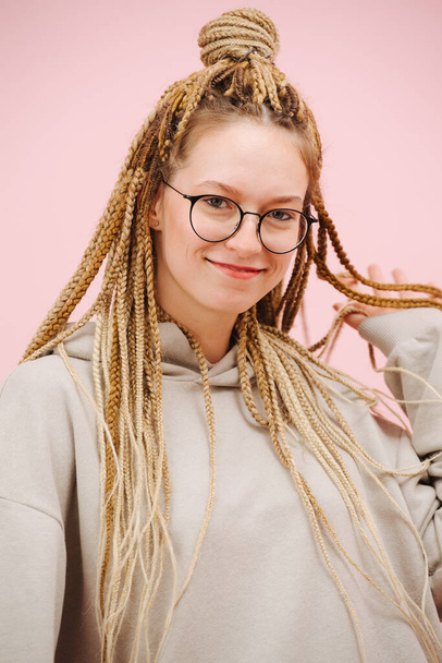 Beautiful smiling woman in round glasses with stylish blond afro braids over pink background. Playing cute, holding hair in hand. - Photo, Image