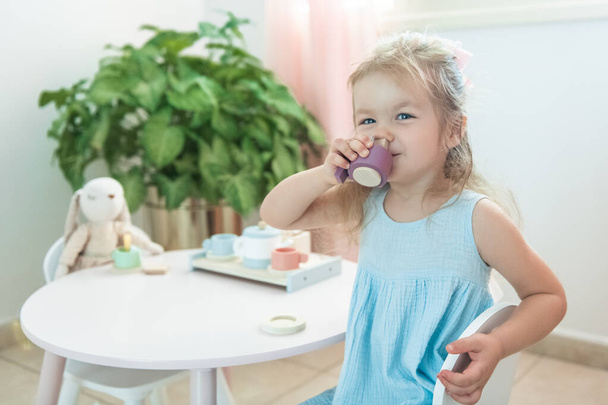 Toddler fair haoired girl is drinking role play tea with rabbit sitting on the wooden chair in blue dress - Φωτογραφία, εικόνα