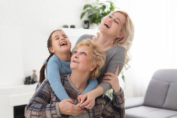 Three generations of women. Beautiful woman and teenage girl are kissing their granny while sitting on couch at home - Photo, image