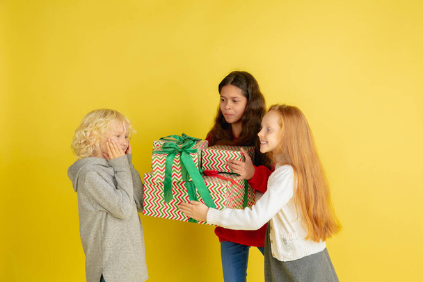 Giving and getting presents on Christmas holidays. Group of happy smiling children having fun isolated on yellow studio background - Photo, Image