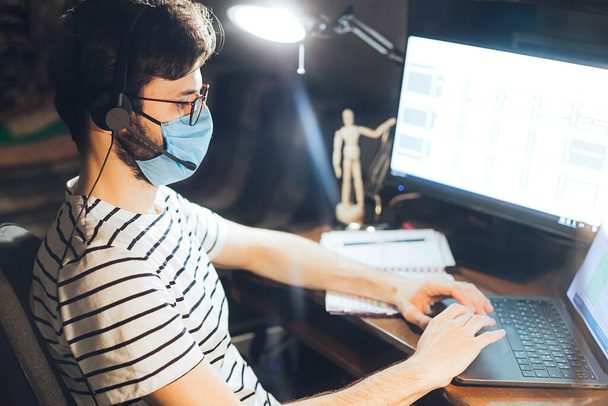 Un hombre caucásico adulto con una camisa azul sentado en casa trabajando en un ordenador portátil con una máscara facial protectora durante la enfermedad epidémica del virus de cuarentena - Foto, imagen