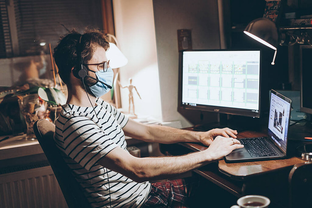 An adult caucasian man wearing a blue shirt sitting at home working on laptop computer wearing a protective face mask during quarantine virus epidemic disease - 写真・画像