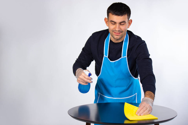 Young handsome man in rubber gloves wipes the table with a rag and homemade spray. Gender stereotypes. Photo on white background. High quality photo - Фото, зображення