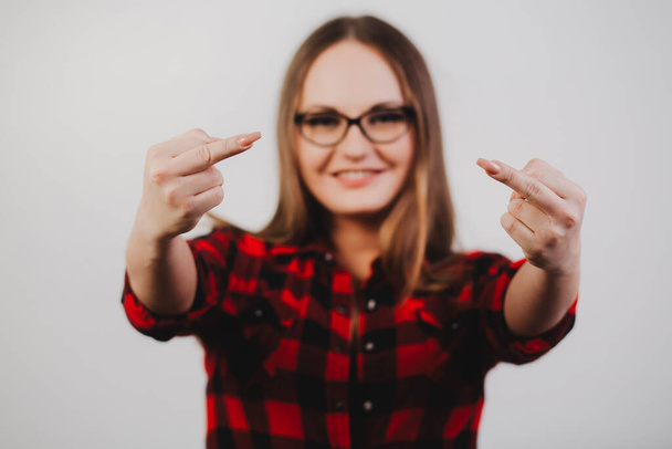 Retrato de uma adolescente de óculos e uma camisa quadriculada em um fundo branco mostrando o dedo médio. - Foto, Imagem