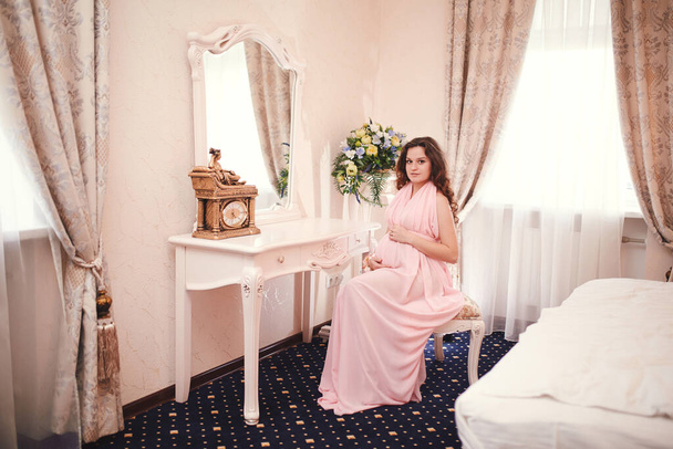 ciężarna rudowłosa kobieta w różowej sukience, siedząca na krześle w klasycznym wnętrzu przy oknie. Czekam na dziecko. ciąża. - Zdjęcie, obraz