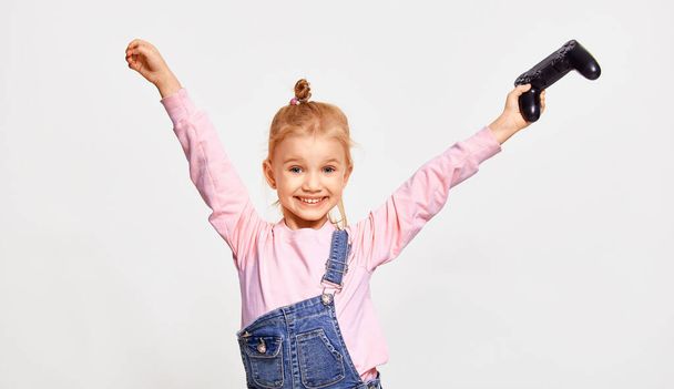 Cute kid girl playing video games holding gamepad and smiling , wearing pink casual sweatshirt and jeans jumpsuit, on white background - Photo, image