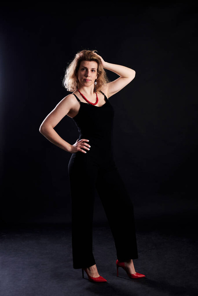 Young blond curly woman, posing on black background. Female dark key photography with in photo studio with lights.Full-length portrait of female, wearing black clothes and red high heels.Love yourself - Фото, изображение