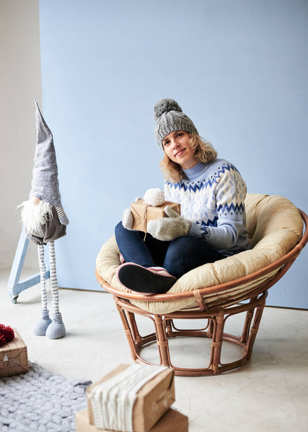 Young woman, wearing light blue sweater with norwegian swedish pattern,grey hat and mittens, is about to open present, sitting in comfy chair in light decorated room.Merry Christmas and Happy new year - 写真・画像