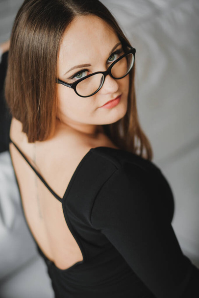 πορτρέτο ενός νεαρού κοριτσιού με γυαλιά και ένα μαύρο φόρεμα που κάθεται σε ένα λευκό καναπέ. - Φωτογραφία, εικόνα