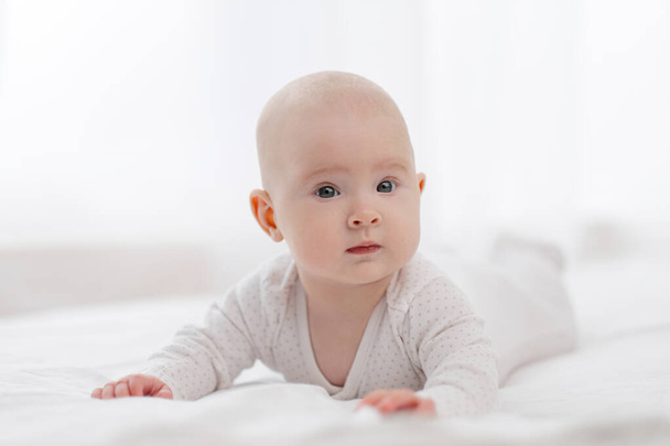 Portrait of a cute beautiful baby with gray eyes in white pajamas on the bed against the window. - Foto, Imagen