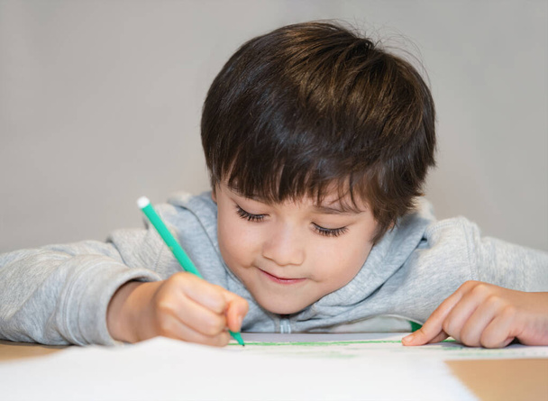 Őszinte lövés boldog gyerek ül egyedül csinál házi feladatot, Gyermek fiú füstölgő arc kezében zöld toll rajz és színezés fehér papíron, Általános iskola és magániskola koncepció - Fotó, kép