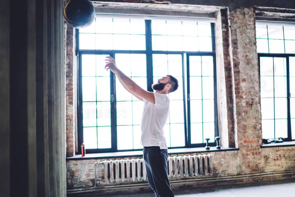 Vue latérale du bel homme barbu en vêtements de sport jouant au basket-ball et sautant avec les mains levées tout en s'entraînant dans une salle de gym spacieuse légère - Photo, image