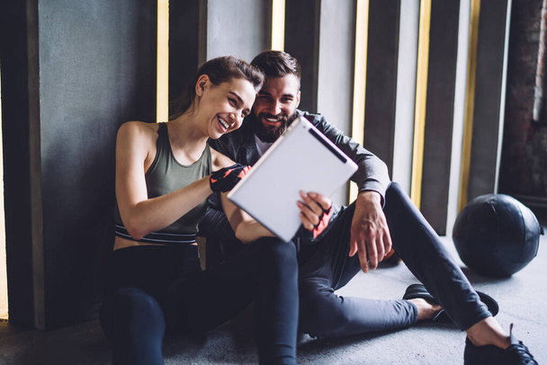 Young positive caucasian couple in love watching videos on tablet together resting after training in gym, smiling sportsman and woman using touchpad for share content about exercising and slimming - Zdjęcie, obraz