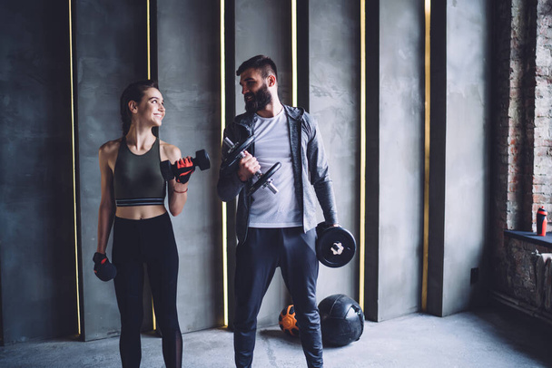 Slim sportswoman and bearded trainer in activewear standing near gray wall and looking at each other while doing dumbbells curls in modern gym - Foto, Imagen