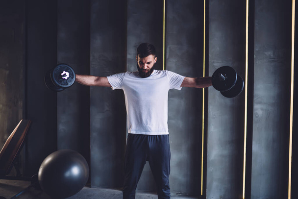 Serious athlete in white shirt with effort lifting weights and experiencing stress during training in modern gym and looking down - Foto, afbeelding