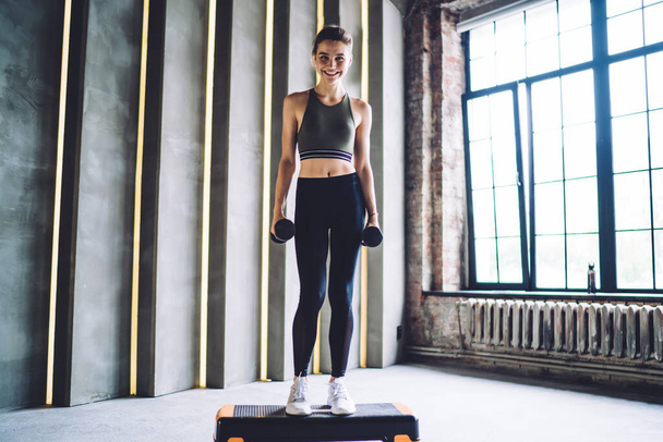 Full body of slim happy female in activewear standing on step platform with dumbbells and looking at camera during workout - Foto, immagini