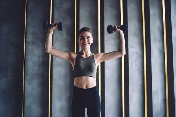 Powerful young female in sports bra raising arms and smiling while doing exercise with dumbbells during functional workout in gym - Valokuva, kuva