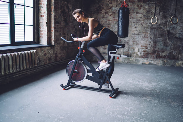 Cheerful caucasian female in earphones enjoying exercising on modern bike in gym having workout, 20s smiling pretty woman satisfied with sound accessory during training losing weight and slimming - Photo, Image