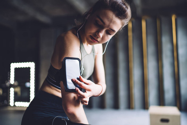 Slim female athlete in sportswear standing in gym and browsing smartphone in armband while listening to music in earphones during workout - Photo, Image