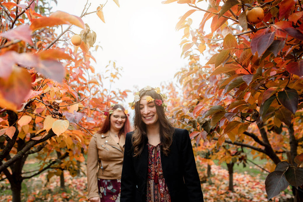 Fehér mosolygós lányok portréja sétál a területen őszi szezonban. Divat, divatos nők virágkoronát viselnek. Vörös levelek az őszi tájban. - Fotó, kép