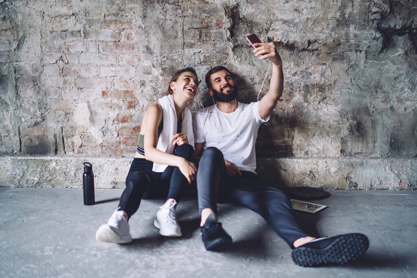 Positive young slim woman and bearded man in sportswear sitting on concrete floor and taking selfie on smartphone after workout - Fotó, kép