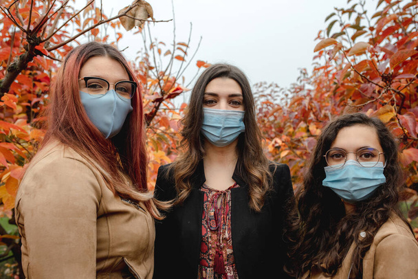 Three girls smiling portrait looking at the camera in a field during fall season. Women wearing protective face mask during coronavirus pandemic. Togetherness, Autumn season, friendship, social distancing concept. - Fotoğraf, Görsel