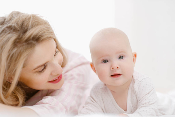 Giovane bella mamma bionda caucasica si trova sul letto con la sua bambina. Luce bianca interno della casa. - Foto, immagini