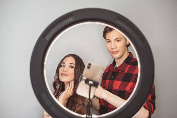 若いカップルの美容ブロガー、ヘアスタイリスト、美容師とモデルとともにヘアスタイル撮影携帯電話でリングランプグレーの壁の背景に - 写真・画像