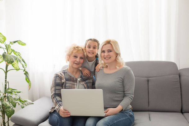 Happy three generations of women sit relax on couch laugh watching funny video on laptop, smiling positive females grandmother, mother and daughter have fun rest on sofa enjoy movie on computer - Photo, Image