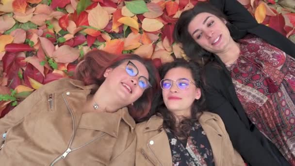 Top view three girls lying down a fallen leaves blanket with smiling faces with during Autumn season. Red, yellow color, fun, friendship concept. - Footage, Video