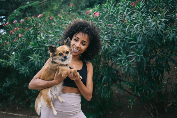 Happy young African American lady with curly hair in sport outfit hugging adorable little chihuahua against green plants in garden - 写真・画像