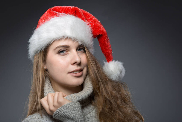 retrato de una bonita chica navideña con sombrero de santa claus con cuello alto delante de un fondo gris oscuro - Foto, imagen